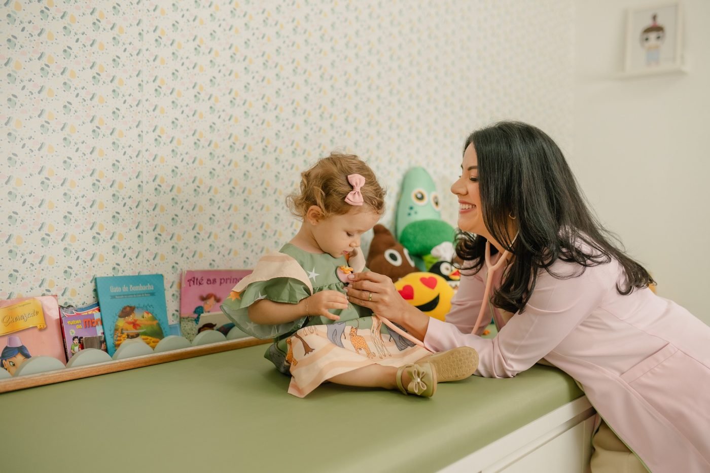 Dra. Raquel Vargas brincando com uma bebê sentada na maca do consultório, cercada de livros e bichos de pelúcia